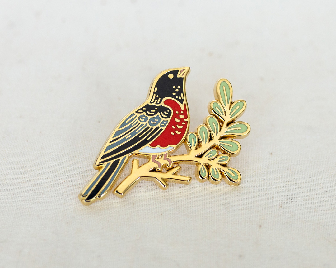 an enamel pin of a robin on a leafy branch in gold metal