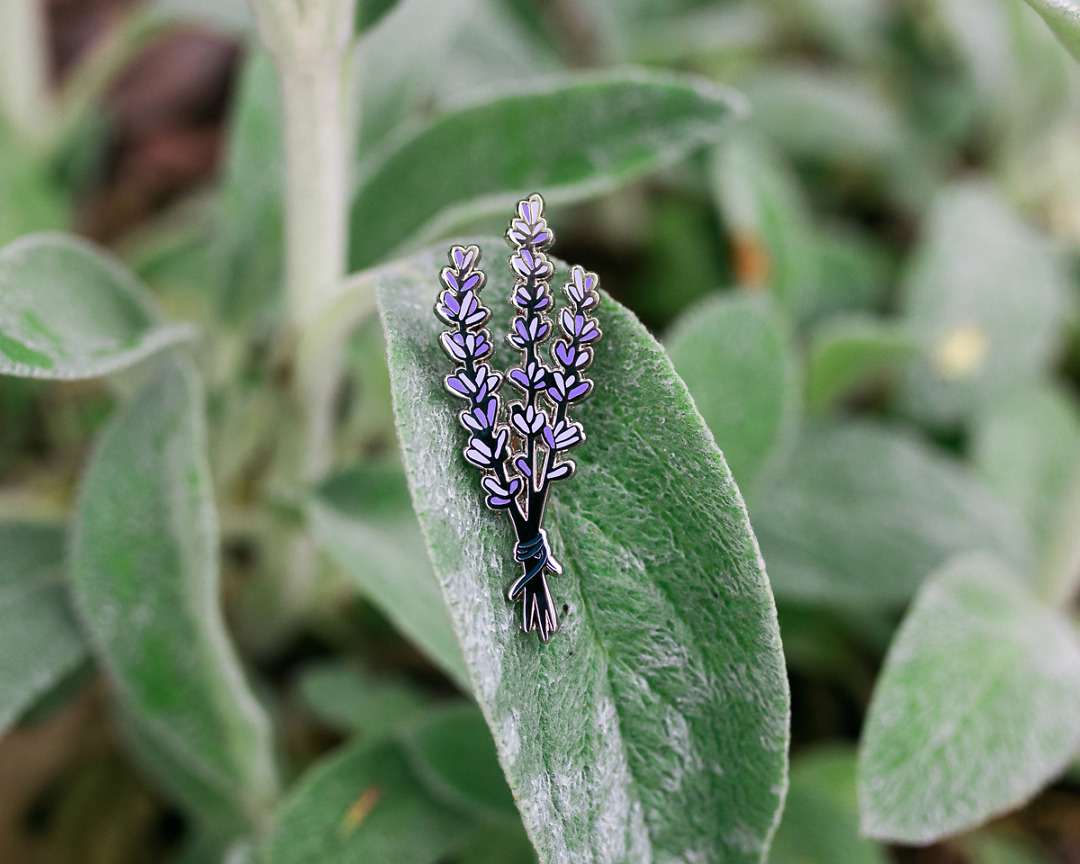 a pretty lavender enamel pin against a fuzzy leaf outside