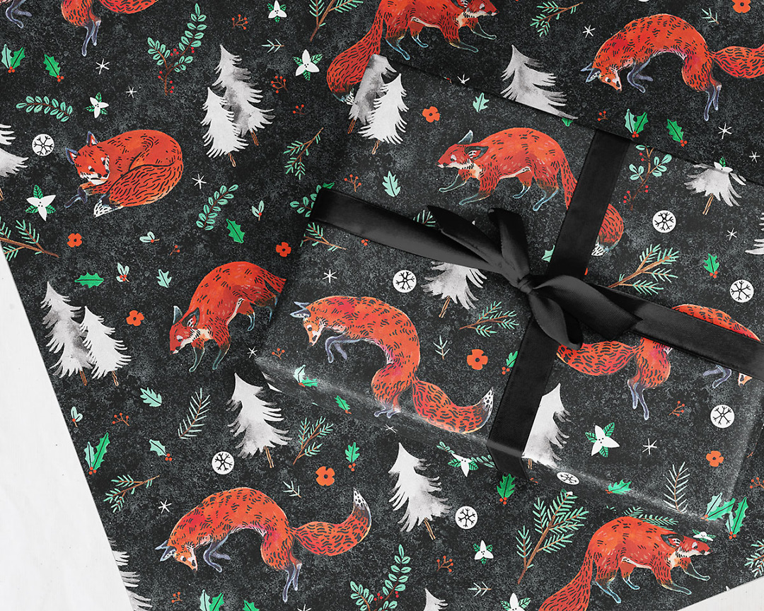 wrapped gift on top of wrapping paper sheet of Wildship Studio holiday gift wrap with fox, pine trees, snowflakes, poinsettia, and holly