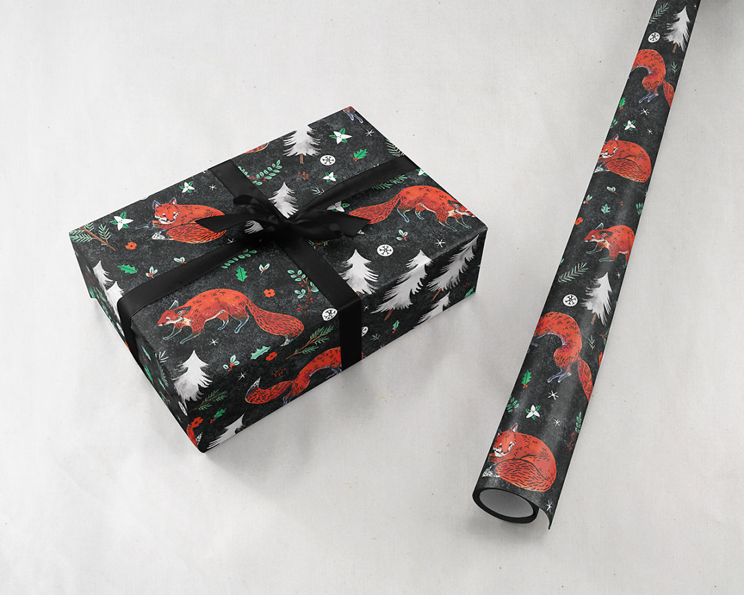 present tied with a black ribbon next to a roll of Wildship Studio holiday gift wrapping paper with fox, pine trees, snowflakes, poinsettia, and holly